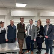 At the school kitchen at Ysgol Calon Cymru’s Llandrindod Well campus, Powys County Council’s Cabinet Member for a Connected Powys, Cllr Jake Berriman (third from left) shakes hands with Matt Lewis, Managing Director at Castell Howell