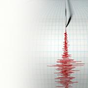 A closeup of a seismograph machine needle drawing a red line on graph paper depicting seismic and earthquake activity.
